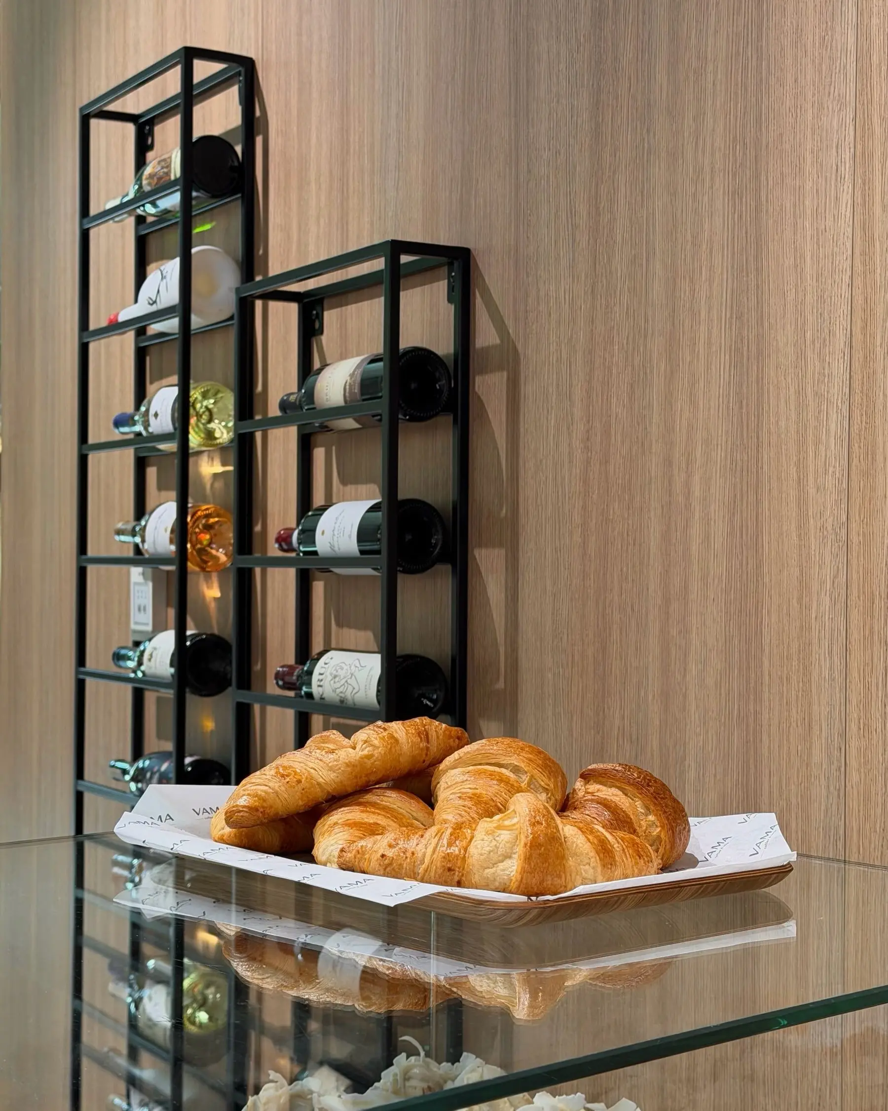 Schwarzes Weinregal mit Flaschen vor der Holzwand, davor ein Korb Croissants