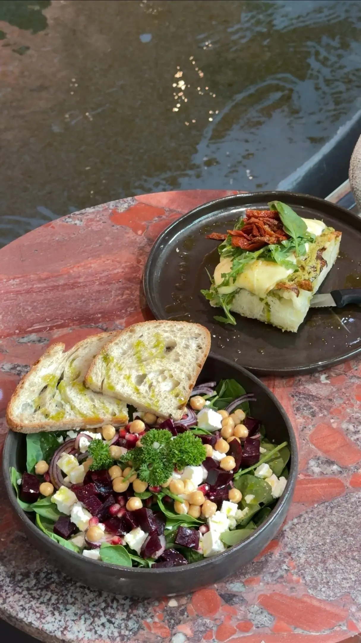 Belegte Focaccia, Brot mit Olivenöl und eine bunte Bowl auf dem roten Steintisch